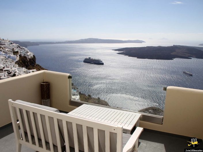 Un balcon avec une table et des bancs blancs surplombe un vaste plan d’eau avec des îles et un navire lointain, sous un ciel bleu clair. Des bâtiments blancs sont perchés sur le flanc gauche, à Santorin en Grèce.