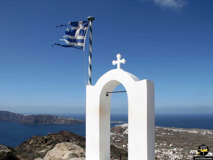 Une arche blanche avec une croix et un drapeau grec au sommet d'une colline, surplombant le paysage côtier de Santorin en Grèce, avec des bâtiments et la mer en arrière-plan.