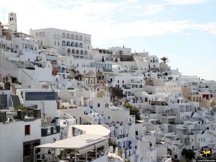 Bâtiments blancs aux accents bleus à flanc de colline sous un ciel bleu, représentant l'architecture à Santorin en Grèce.