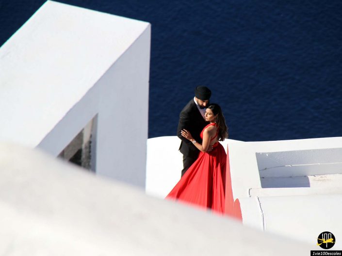 Un couple pose sur un toit blanc à Santorin en Grèce, surplombant une mer d'un bleu profond. La femme porte une robe rouge fluide et l'homme est vêtu d'un costume noir et d'un chapeau.
