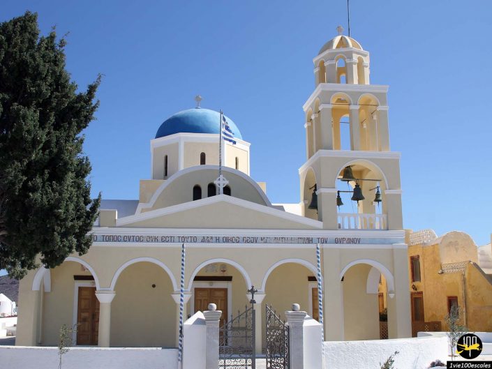 Une église avec un dôme bleu et un clocher, doté de portes et de fenêtres cintrées, se dresse sous un ciel bleu clair, avec un arbre sur le côté gauche, à Santorin en Grèce.