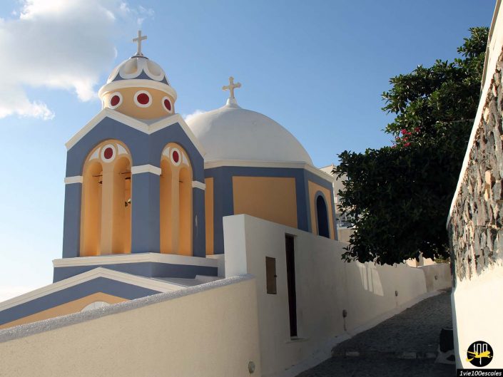 Une église traditionnelle bleue et blanche avec des dômes et des croix se dresse le long d'un chemin étroit dans une ville côtière ensoleillée, à Santorin en Grèce.