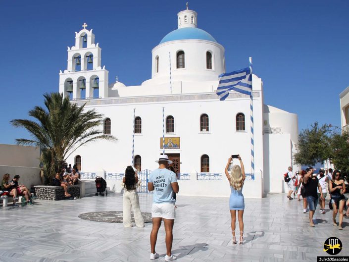 Un groupe de personnes, dont un couple prenant des photos, se tiennent devant une église blanche avec un dôme bleu et un clocher sur une place ensoleillée à Santorin en Grèce. Deux drapeaux grecs flottent et un palmier se trouve à proximité.