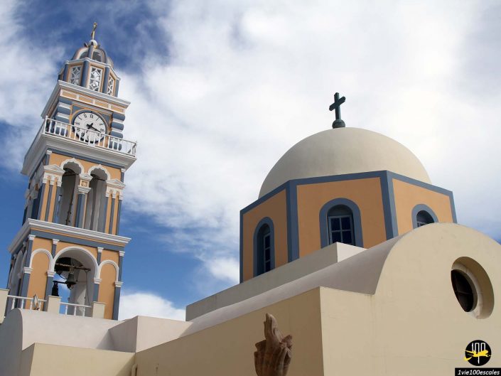 Une église avec une tour d'horloge bleu clair et un dôme avec une croix sur un ciel partiellement nuageux, à Santorin en Grèce.