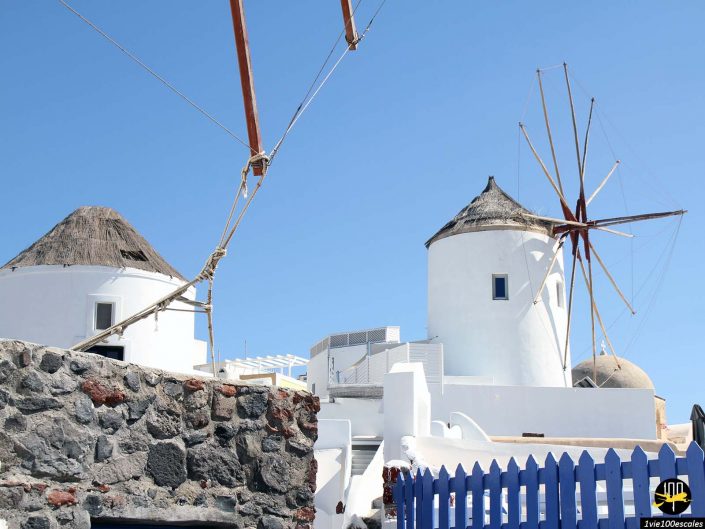 Vue de deux moulins à vent blancs aux toits de chaume dans une zone lumineuse et ensoleillée à Santorin en Grèce. Un mur de pierre et une clôture bleue sont également visibles au premier plan. Le ciel est clair et bleu.