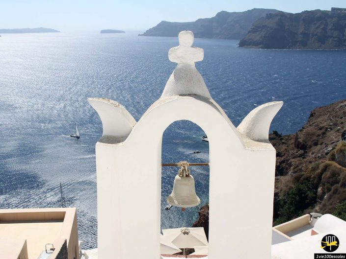 Un clocher blanc se dresse au premier plan surplombant une vaste mer bleue avec des voiliers et des îles vallonnées en arrière-plan, à Santorin en Grèce.