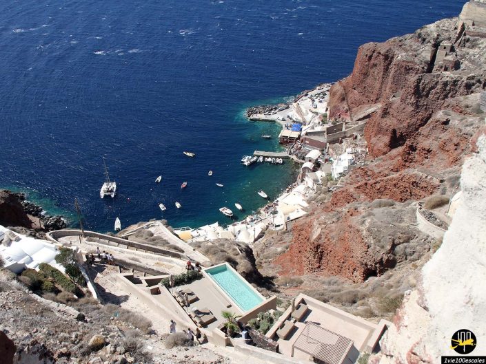 Vue depuis une falaise à Santorin en Grèce surplombant un village balnéaire avec des bâtiments blancs, des bateaux dans l'eau bleue et une piscine au premier plan. Le terrain rocheux et escarpé mène à la petite zone portuaire.