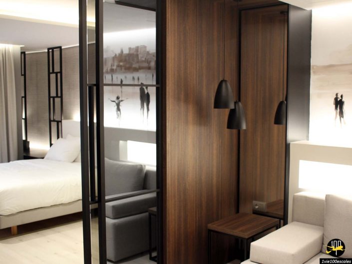 Chambre d'hôtel moderne à Athènes en Grèce avec un lit, un canapé et des touches de bois sombre. La pièce présente une décoration minimaliste avec des œuvres d'art murales en niveaux de gris et deux lampes noires suspendues.