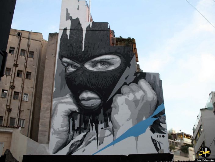 Une grande fresque murale à Athènes en Grèce représente une personne avec un masque noir couvrant son visage, seuls ses yeux et sa bouche étant visibles. L’œuvre présente des tons gris et blancs avec un trait bleu audacieux.