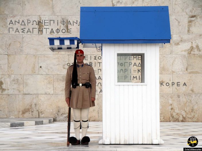 Une garde de cérémonie en uniforme traditionnel se tient à côté d'un petit corps de garde blanc et bleu, à Athènes en Grèce, devant un mur de pierre aux inscriptions grecques.