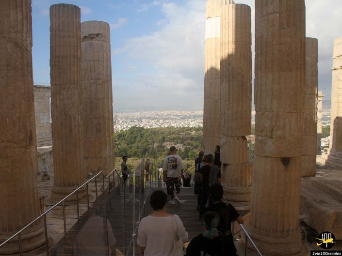 Les gens marchent à travers des colonnes anciennes surplombant un paysage urbain et de la verdure en contrebas, avec un ciel clair en arrière-plan, à Athènes en Grèce.