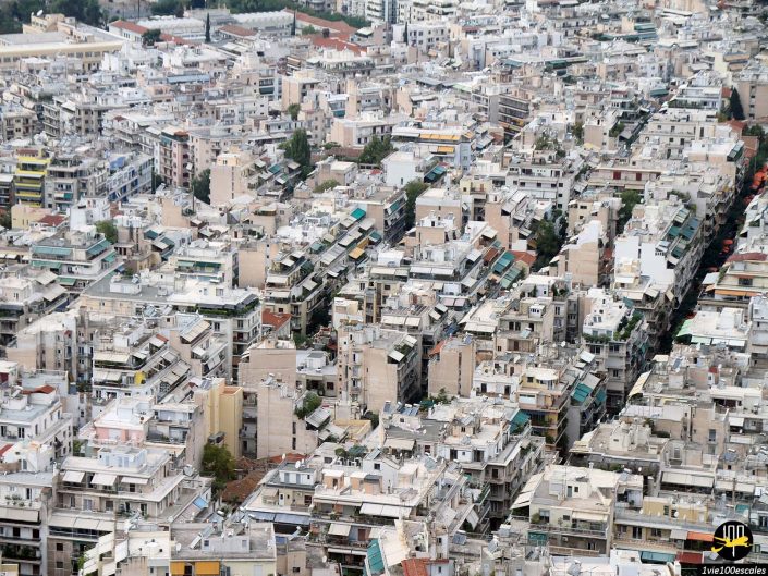 Vue aérienne d'une zone urbaine densément peuplée à Athènes en Grèce avec de nombreux bâtiments à plusieurs étages, certains avec des structures sur le toit et de la verdure, donnant l'impression d'un quartier urbain animé.