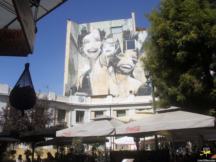 Fresque de rue représentant trois visages souriants sur un grand bâtiment au-dessus des parasols d'un café en plein air dans une zone ensoleillée et arborée, à Athènes en Grèce.