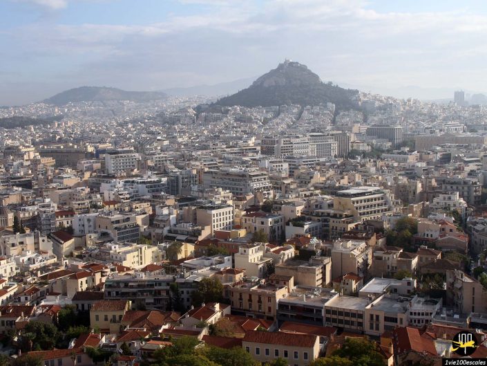 Une vue panoramique d'un paysage urbain densément peuplé à Athènes en Grèce, avec une montagne surmontée de bâtiments au loin, sous un ciel partiellement nuageux.