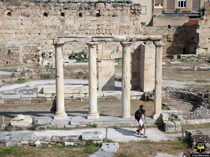 Deux touristes se promènent près de ruines antiques avec de hautes colonnes de pierre et diverses structures, situées dans une zone largement ouverte et partiellement érodée. Un haut mur de pierre avec quelques bâtiments modernes se trouve en arrière-plan, à Athènes en Grèce.