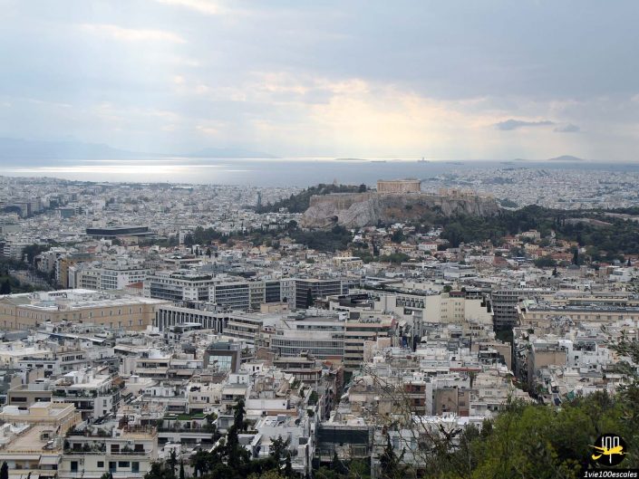 Une vue panoramique d'une ville avec de nombreux bâtiments, un site historique important au sommet d'une colline et un plan d'eau au loin sous un ciel nuageux à Athènes en Grèce.