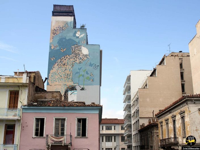 Un paysage urbain à Athènes en Grèce présente une grande fresque murale représentant une personne avec des plantes et des papillons sur le côté d'un bâtiment à plusieurs étages. Les bâtiments environnants semblent usés et vieillis.