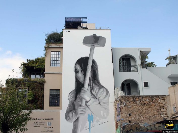Une grande fresque peinte sur le côté d'un bâtiment blanc à Athènes en Grèce représente une personne tenant une masse. Les bâtiments qui l’entourent sont recouverts de verdure et une partie du mur présente des graffitis plus petits.