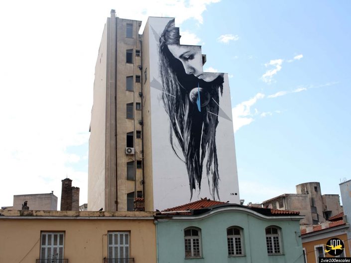 Une grande fresque murale sur le côté d'un grand immeuble à Athènes en Grèce représente une femme aux cheveux longs, la tête légèrement inclinée vers le bas et une petite sphère lumineuse devant son visage. La fresque murale s'intègre parfaitement à la structure du bâtiment.