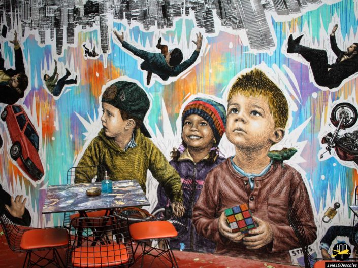 Murale colorée représentant trois enfants, dont un tenant un Rubik's Cube. Autour d’eux, des gens, des voitures et des motos semblent tomber d’un paysage urbain. Des chaises rouges et une table sont au premier plan à Athènes en Grèce.