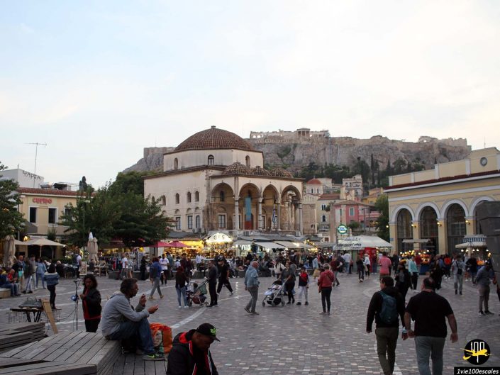 Les gens se rassemblent et se promènent sur la place Monastiraki à Athènes en Grèce, avec l'Acropole visible en arrière-plan et divers commerces et bâtiments entourant le quartier.