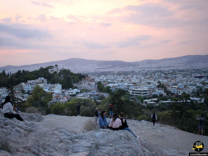 Un groupe de personnes est assis sur un terrain rocheux surplombant une ville avec des bâtiments dispersés et des montagnes au loin sous un ciel nuageux, à Athènes en Grèce.