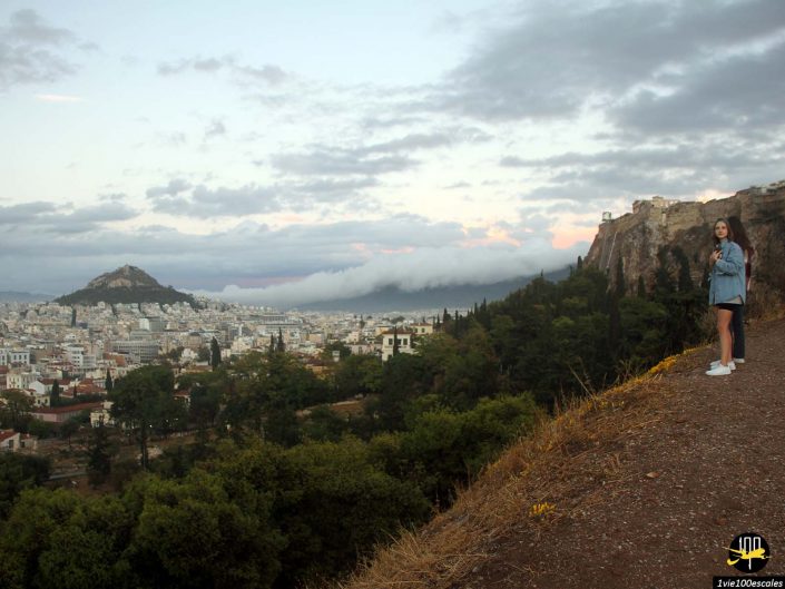 Une femme se tient sur une colline surplombant un paysage urbain avec des bâtiments et de la verdure, à Athènes en Grèce. En arrière-plan, une autre colline avec des structures est visible sous le ciel nuageux.