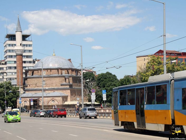 Une scène de rue à Sofia, en Bulgarie, montre une vieille mosquée avec un grand minaret, des bâtiments modernes en arrière-plan, des voitures garées le long de la route et un tramway jaune et bleu qui passe.