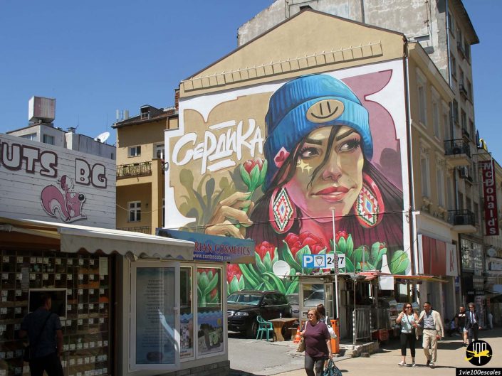 Une grande fresque murale sur le côté d'un immeuble à Sofia en Bulgarie représente une femme portant un bonnet bleu avec un visage souriant. Ci-dessous se trouvent divers magasins et personnes marchant.