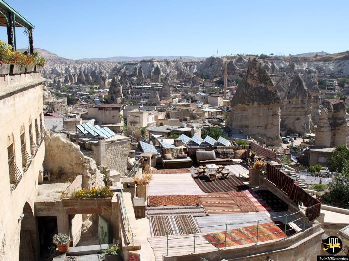 Une vue de la Cappadoce, en Turquie, avec des formations rocheuses et un toit-terrasse orné de tapis et de coussins traditionnels. Le paysage comprend des maisons intégrées aux structures rocheuses.