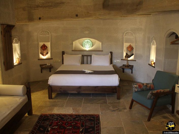Une chambre confortable en Cappadoce en Turquie avec un lit en bois, une literie blanche et des murs en pierre dotés d'étagères intégrées avec des décorations. Un fauteuil, un canapé et un petit tapis complètent le décor rustique de la pièce.