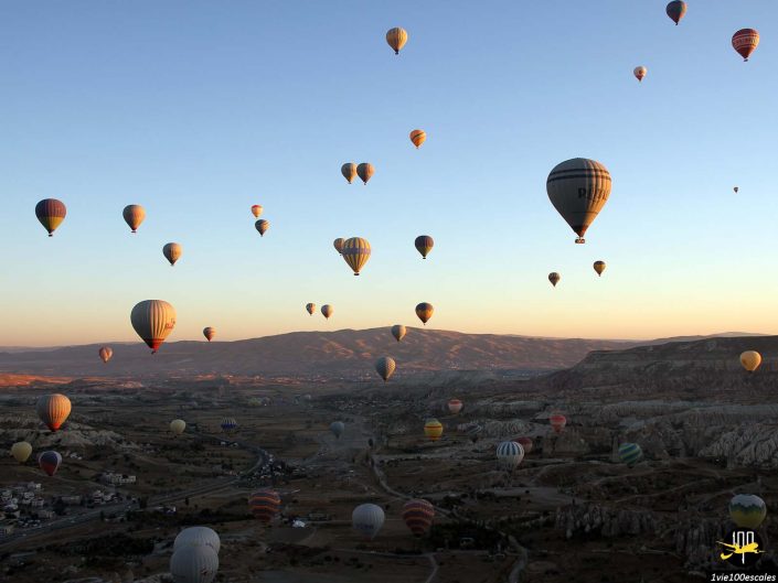 Un grand nombre de montgolfières flottent dans le ciel au-dessus d'un vaste paysage vallonné au coucher du soleil en Cappadoce en Turquie.