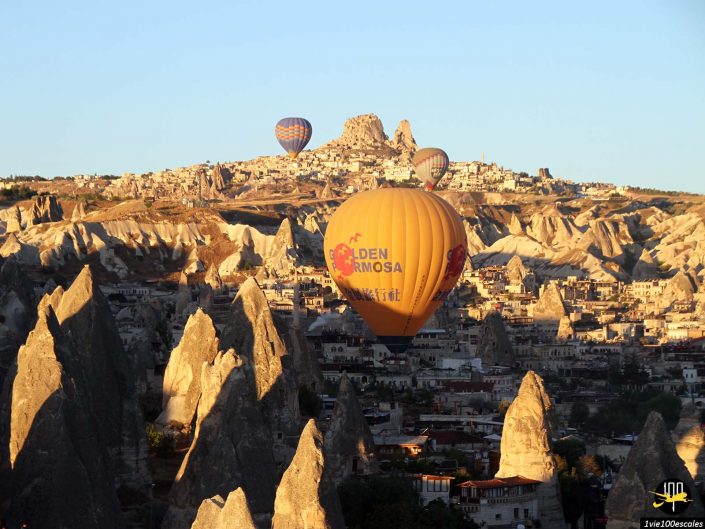 Montgolfières survolant un paysage rocheux avec des formations rocheuses uniques et une ville en arrière-plan sous un ciel clair, en Cappadoce en Turquie.