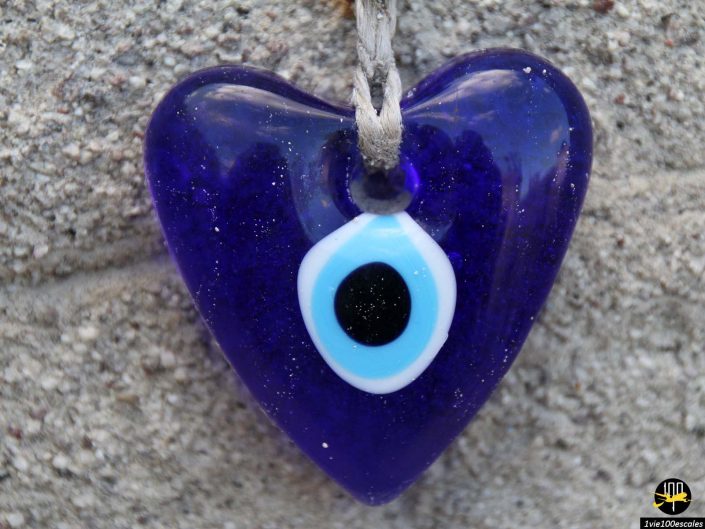 Un pendentif en verre bleu en forme de cœur avec un symbole d'œil au centre, suspendu sur un fond de pierre brute, rappelant les trésors trouvés en Cappadoce en Turquie.