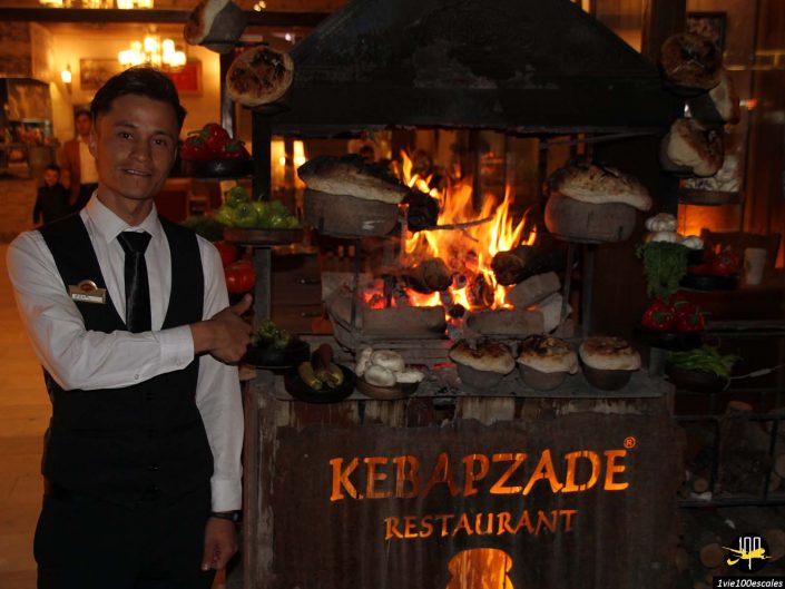 Un serveur en gilet noir et cravate se tient à côté du grill d'un restaurant rempli de divers aliments cuisant sur une flamme nue, avec une pancarte indiquant "Kebapzade Restaurant" visible en dessous. Cette scène se déroule en Cappadoce en Turquie, ajoutant une touche authentique à l'expérience culinaire.