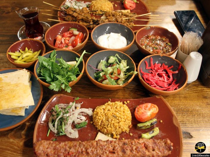 Une variété de plats du Moyen-Orient, rappelant les riches traditions culinaires de Cappadoce en Turquie, notamment des brochettes de viande, des légumes, du pain, des sauces et des thés, sont disposés sur une table en bois.