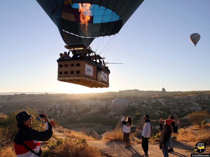 Une montgolfière avec des passagers survole un paysage pittoresque au lever du soleil en Cappadoce en Turquie. Des personnes au sol photographient le ballon tandis qu'un autre ballon flotte en arrière-plan.