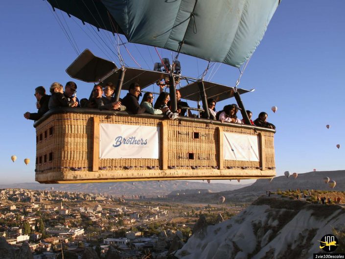 Un groupe de personnes dans une nacelle de montgolfière en osier étiquetée « Frères » survole un paysage pittoresque en Cappadoce en Turquie, avec d'autres montgolfières parsemant le ciel et une charmante ville en contrebas.