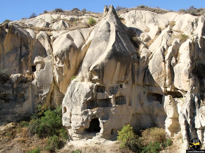 Formations rocheuses avec des habitations troglodytes dans un paysage accidenté, comportant un rocher central pointu et diverses ouvertures et fenêtres creusées dans la paroi rocheuse, en Cappadoce en Turquie. Des buissons et une végétation clairsemée entourent la zone.