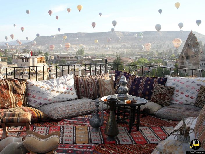 Coin salon extérieur avec coussins et tapis à motifs donnant sur un paysage avec de nombreuses montgolfières dans le ciel en Cappadoce en Turquie.