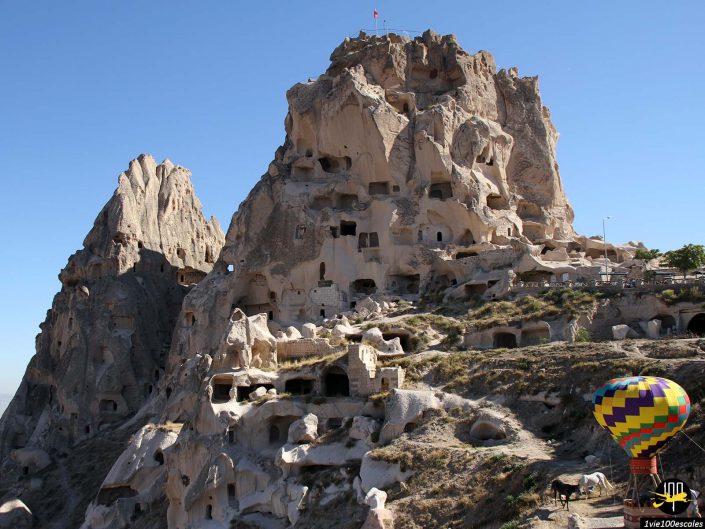 Colline rocheuse avec de nombreuses habitations troglodytes en Cappadoce en Turquie, une montgolfière vibrante au premier plan et un ciel bleu clair.