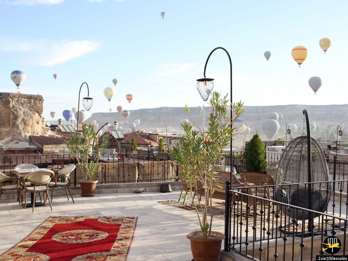 Un toit-terrasse doté de tables, de chaises et de plantes en pot surplombe un paysage pittoresque de Cappadoce en Turquie, où de nombreuses montgolfières remplissent le ciel. Les montagnes et le ciel dégagé offrent une toile de fond époustouflante.