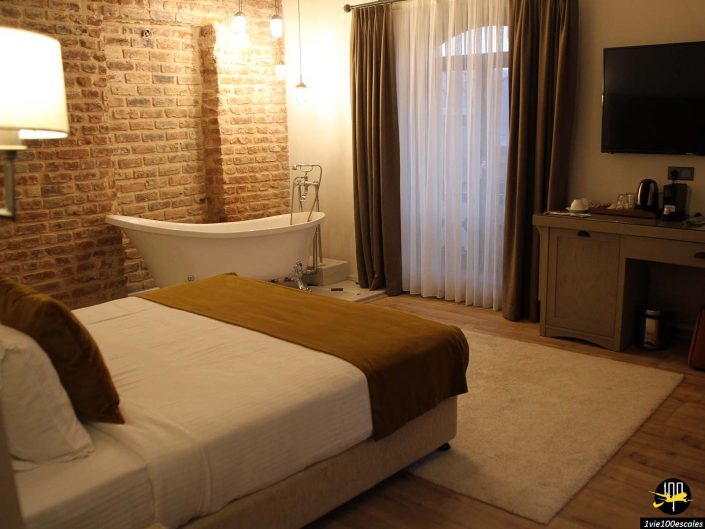 Chambre d'hôtel avec lit double, mur de briques apparentes, baignoire îlot, télévision murale, bureau et grands rideaux recouvrant une fenêtre à Istanbul en Turquie.