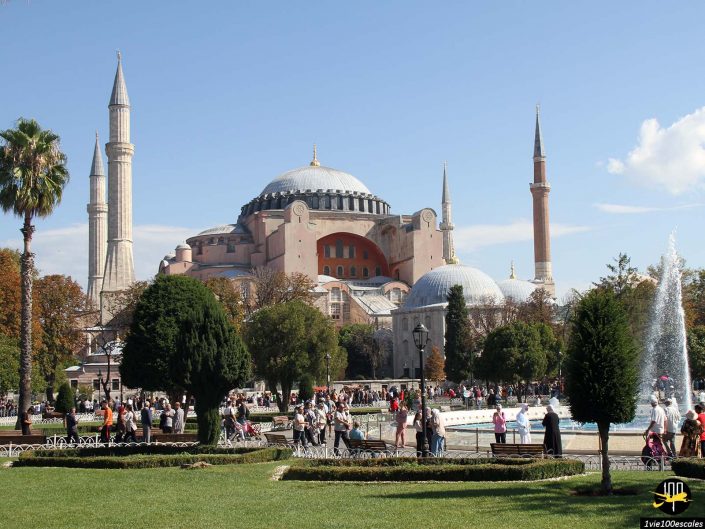 Un grand bâtiment historique avec des dômes et des minarets se dresse entouré d'arbres, à Istanbul en Turquie, avec un parc rempli de monde. Une fontaine est visible à droite.