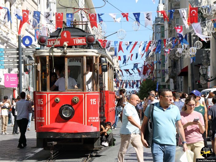 Un tramway rouge du patrimoine circule dans une rue animée d'Istanbul en Turquie, ornée de drapeaux et de décorations, avec des piétons marchant des deux côtés.