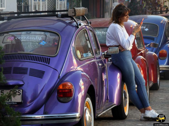 Une femme portant des lunettes de soleil s'appuie contre une voiture classique violette, utilisant son smartphone à Istanbul en Turquie. D'autres voitures classiques, dont une rouge, sont garées à proximité dans la rue pavée.