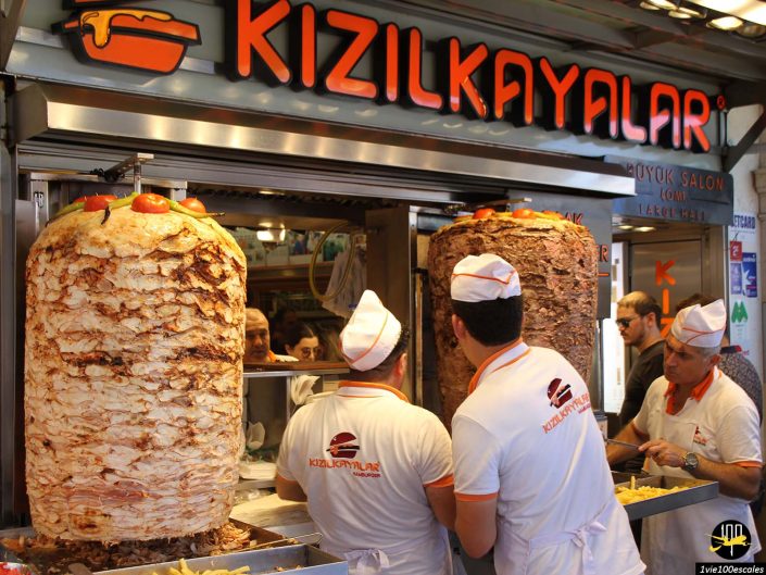 Trois ouvriers en uniforme blanc préparent la nourriture dans un restaurant Kizilkayalar à Istanbul en Turquie, coupant de grandes rôtissoires verticales de viande de doner kebab. Deux clients attendent devant le comptoir.