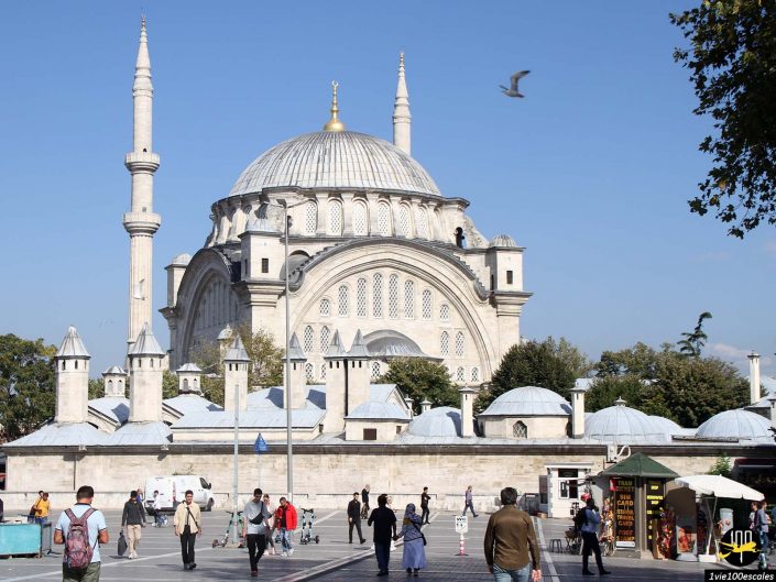 Les gens marchent devant la mosquée Yeni, une mosquée historique avec deux minarets et un grand dôme, sous un ciel bleu clair à Istanbul en Turquie ; un oiseau vole au-dessus.