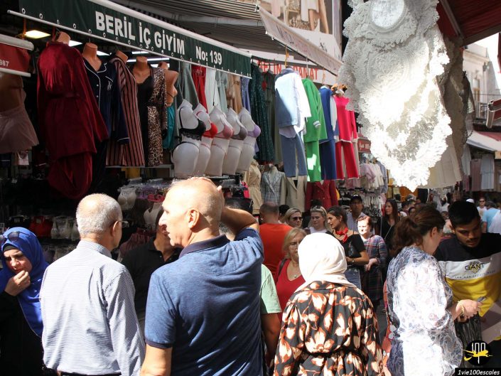 Un marché extérieur bondé à Istanbul en Turquie, avec des gens parcourant les étals vendant divers vêtements, notamment de la lingerie, des chemises et des vestes. Un grand tissu en dentelle est bien en vue au premier plan.