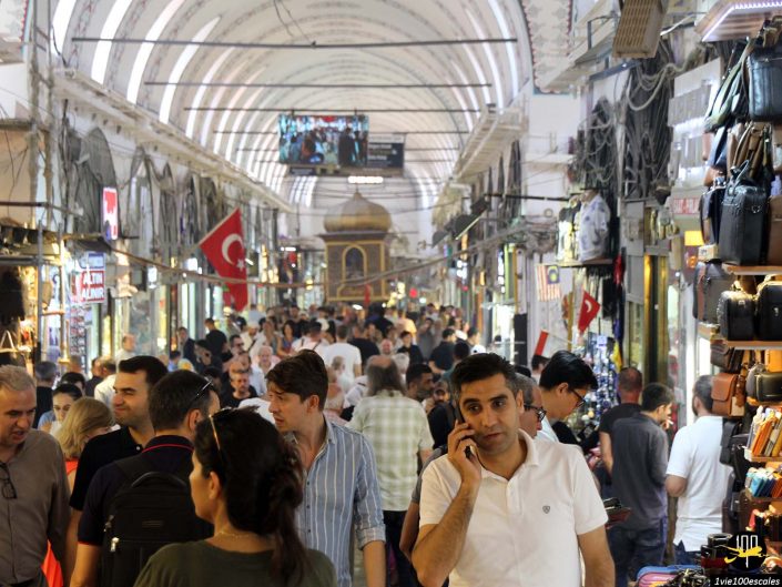 Le marché couvert bondé, à Istanbul en Turquie, regorge de nombreuses personnes marchant et faisant du shopping. Divers magasins bordent les murs et un homme au premier plan parle au téléphone. Le drapeau turc est visible au-dessus de la scène animée.